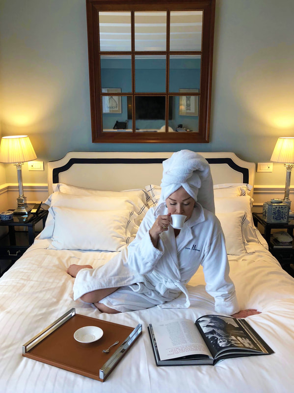Mrs. Stafford in a white robe sipping coffee & reading on a hotel bed