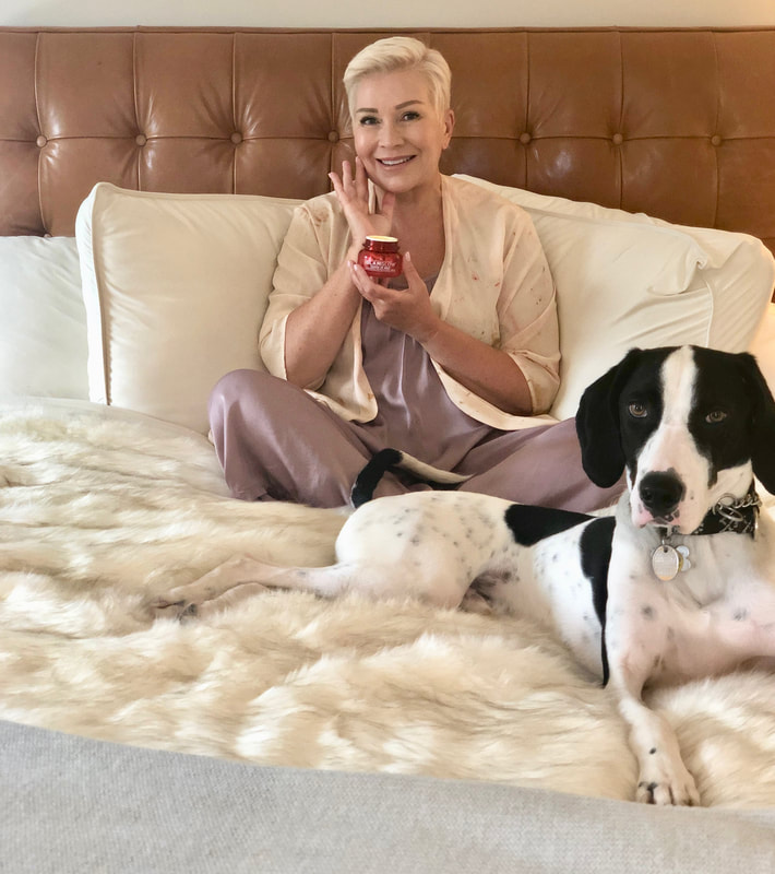 Mrs. Stafford sitting on her bed, with her dog, & holding a skincare product